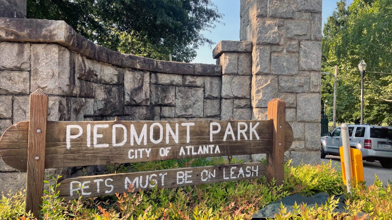 A rustic wooden sign for Piedmont Park in Atlanta, GA, featuring a stone wall background and a clear pets must be on leash note.